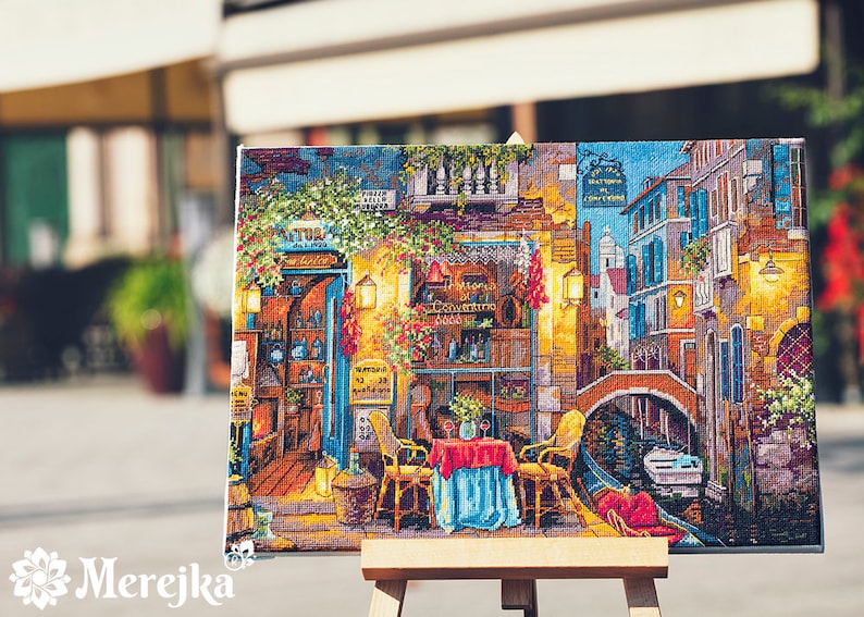 Puede incluir: Una obra de punto de cruz colorida que representa una pintoresca escena de calle italiana con un canal, un puente y un caf&eacute; con mesas y sillas. La obra est&aacute; enmarcada y expuesta en un caballete.