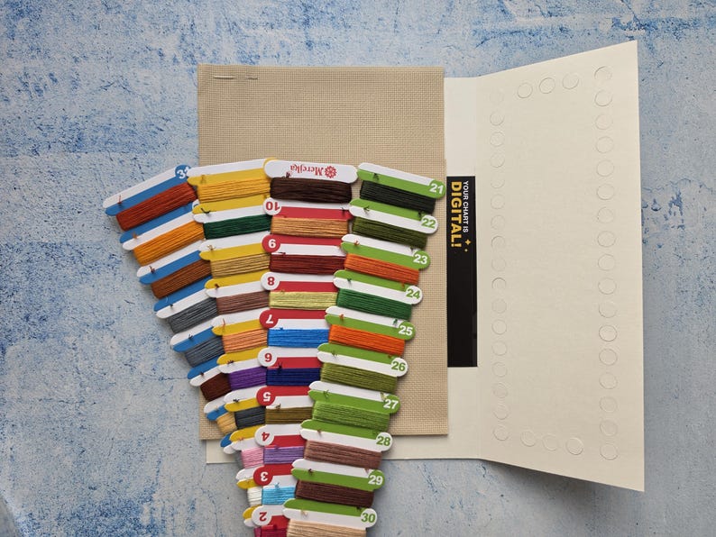 May include: A cross stitch kit featuring a beige Aida fabric square and multiple cardboard thread organizers holding various colored embroidery floss. The organizers are numbered and include a black card with the text YOUR CHART IS DIGITAL!