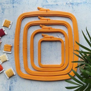 Puede incluir: Cuatro aros de bordado naranja de diferentes tamaños, anidados uno dentro del otro. Los aros están sobre un fondo azul claro con carretes de hilo de bordar de varios colores.