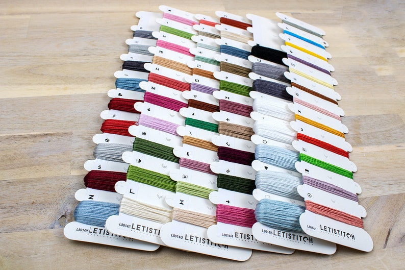 May include: A collection of embroidery floss spools organized by color and labeled with letters. The spools are arranged in rows and are all from the same brand, Letistitch.