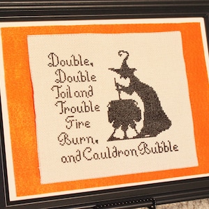 May include: A framed cross-stitch with an orange mount. The cross-stitch features a black silhouette of a witch stirring a cauldron with the text "Double, Double Toil and Trouble Fire Burn, and Cauldron Bubble."