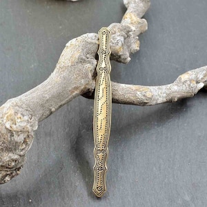 Puede incluir: Una pinza para el pelo de metal dorado con un diseño decorativo. La pinza es larga y estrecha, con un extremo puntiagudo y una parte superior redondeada. El diseño presenta una serie de pequeños círculos y líneas.