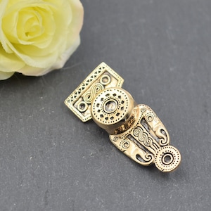 Könnte beinhalten: Goldfarbener Metallanhänger mit aufwendigem keltischem Knotenmuster. Der Anhänger hat ein kreisförmiges Mittelstück mit einer erhabenen, strukturierten Oberfläche.