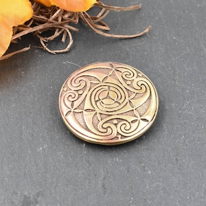 May include: A round, bronze-colored metal object with an intricate Celtic knot design. The design features swirling patterns and a central triquetra. The object is set against a dark gray background, with a hint of orange and brown foliage in the upper left corner.
