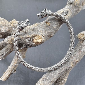 Könnte beinhalten: Ein silbernes geflochtenes Armreif-Armband mit Wolfskopf-Akzenten. Das Armband befindet sich auf einem hölzernen Hintergrund.