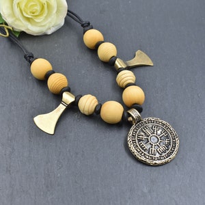May include: A necklace with wooden beads and a metal pendant featuring a circular design with runes. The pendant is attached to a black leather cord.