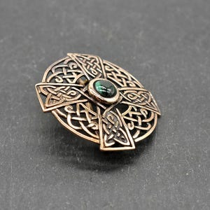 Bronze-farbene keltische Kreuzbrosche mit aufwendigem Knotenmuster. In der Mitte befindet sich ein kleiner, runder, dunkelgrüner Stein. Die Brosche ist vor einem dunkelgrauen Hintergrund platziert, der ihre detaillierte Handwerkskunst zeigt.