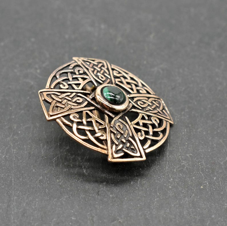 Bronze-farbene keltische Kreuzbrosche mit aufwendigem Knotenmuster. In der Mitte befindet sich ein kleiner, runder, dunkelgrüner Stein. Die Brosche ist vor einem dunkelgrauen Hintergrund platziert, der ihre detaillierte Handwerkskunst zeigt.