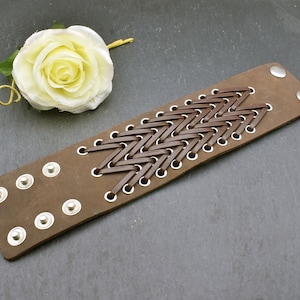 Könnte beinhalten: Ein braunes Lederarmband mit gewebtem Muster und silbernen Metallösen. Das Armband ist verstellbar und hat ein schlichtes, elegantes Design.