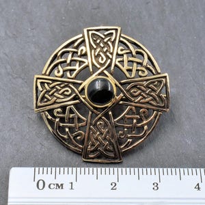Eine goldfarbene keltische Kreuzbrosche mit aufwendigem Knotenmuster. In der Mitte befindet sich ein schwarzer, kreisförmiger Stein. Die Brosche hat einen Durchmesser von etwa 3.5 cm und zeigt detaillierte Handwerkskunst und historische Symbolik.