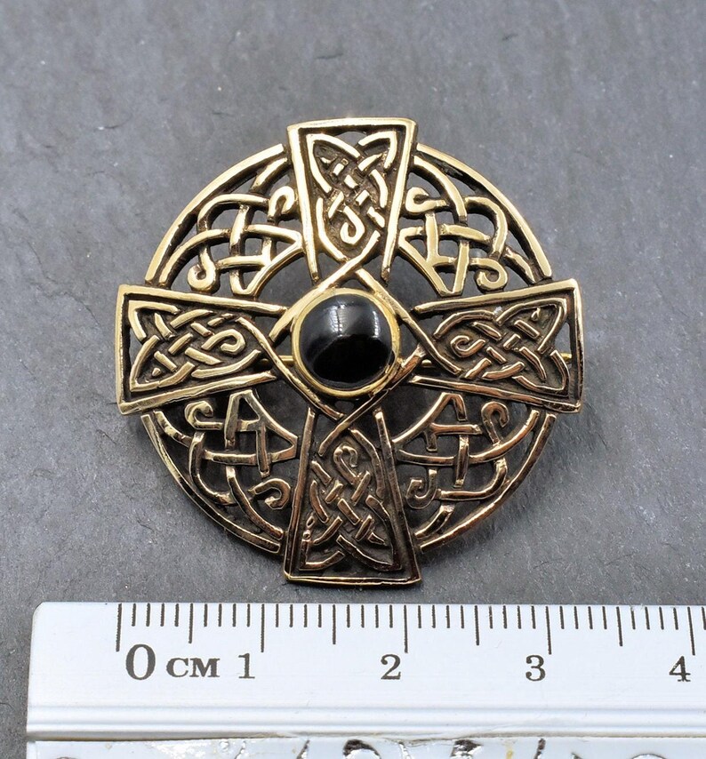 Eine goldfarbene keltische Kreuzbrosche mit aufwendigem Knotenmuster. In der Mitte befindet sich ein schwarzer, kreisförmiger Stein. Die Brosche hat einen Durchmesser von etwa 3.5 cm und zeigt detaillierte Handwerkskunst und historische Symbolik.