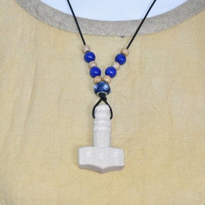 Può includere: Una collana con pendente in legno con un pendente a forma di martello di colore chiaro, infilato su un cordino nero. La collana include perline di legno blu e beige. Il pendente è scolpito con dettagli intricati.