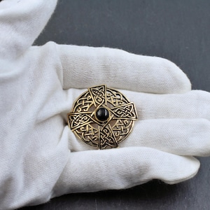 Eine goldfarbene keltische Knotenbrosche mit einem zentralen schwarzen Stein. Die Brosche weist komplizierte Knotenmuster auf und ist kreisförmig. Der Schmuck wird auf einer weißen Oberfläche präsentiert.