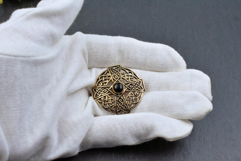 Eine goldfarbene keltische Knotenbrosche mit einem zentralen schwarzen Stein. Die Brosche weist komplizierte Knotenmuster auf und ist kreisförmig. Der Schmuck wird auf einer weißen Oberfläche präsentiert.