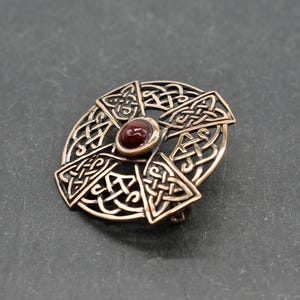 Bronze-farbene keltische Knotenbroschen mit einem zentralen, runden, rotbraunen Stein. Die Brosche hat ein kreisförmiges Design mit Kreuzmotiv, komplizierten Knotenmustern und einem Nadelverschluss auf der Rückseite. Ein dekoratives Accessoire.