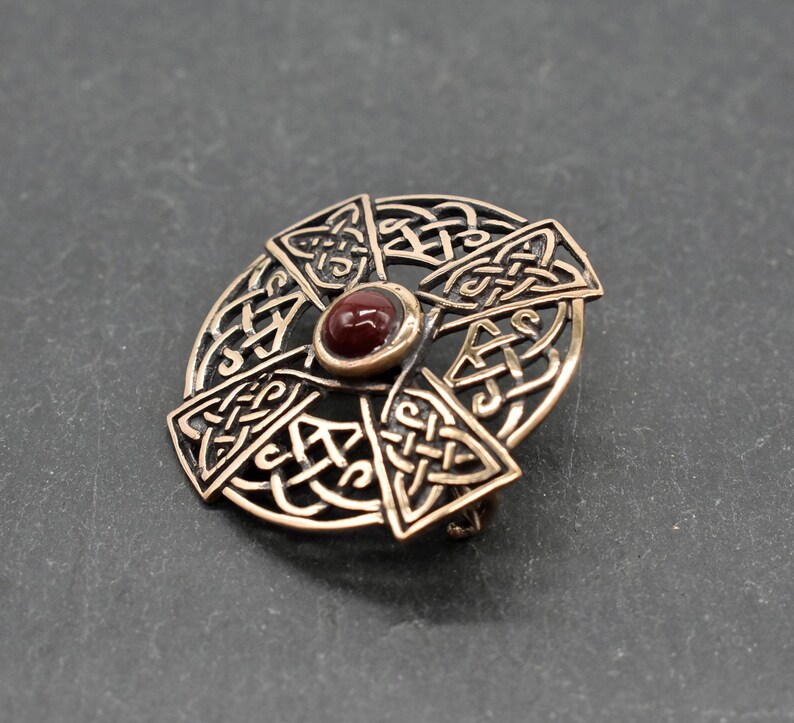 Bronze-farbene keltische Knotenbroschen mit einem zentralen, runden, rotbraunen Stein. Die Brosche hat ein kreisförmiges Design mit Kreuzmotiv, komplizierten Knotenmustern und einem Nadelverschluss auf der Rückseite. Ein dekoratives Accessoire.