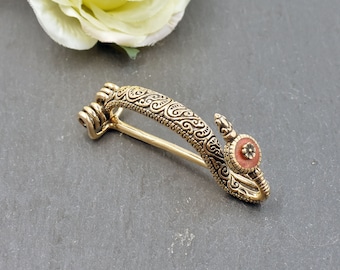 Puede incluir: Un broche dorado en forma de serpiente estilizada con una piedra naranja en el centro de su cabeza. El broche tiene un diseño detallado de patrones en espiral.
