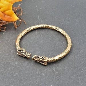 Puede incluir: Un brazalete de metal dorado con un diseño trenzado y dos extremos con cabeza de dragón.