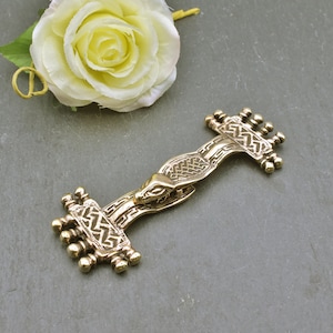 Könnte beinhalten: Eine goldfarbene Metallbrosche mit einem aufwendigen Design, das einen stilisierten Tierkopf und geometrische Muster zeigt. Die Brosche hat eine stangenartige Form mit dekorativen Enden. Eine cremefarbene Rose befindet sich im Hintergrund.