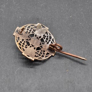 Eine bronzefarbene Brosche im Vintage-Stil mit einem ovalen, durchbrochenen Design. Die Brosche hat einen Nadelverschluss auf der Rückseite. Die filigrane Metallarbeit erzeugt ein zartes, spitzenartiges Muster. Die Brosche hat eine warme, antike Bronze Farbe.