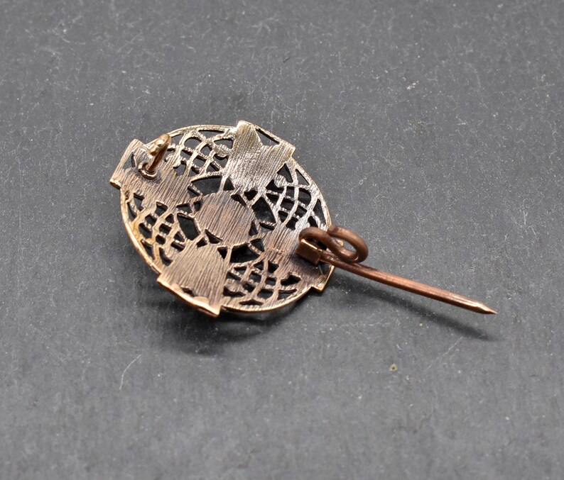 Eine bronzefarbene Brosche im Vintage-Stil mit einem ovalen, durchbrochenen Design. Die Brosche hat einen Nadelverschluss auf der Rückseite. Die filigrane Metallarbeit erzeugt ein zartes, spitzenartiges Muster. Die Brosche hat eine warme, antike Bronze Farbe.