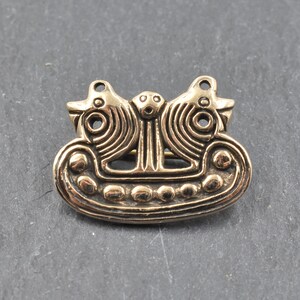 Eine goldfarbene Brosche aus Metall mit einem Design aus zwei stilisierten Vögeln, die sich gegenüberstehen. Die Vögel sitzen auf einem bootförmigen Sockel mit einer Reihe runder Akzente.