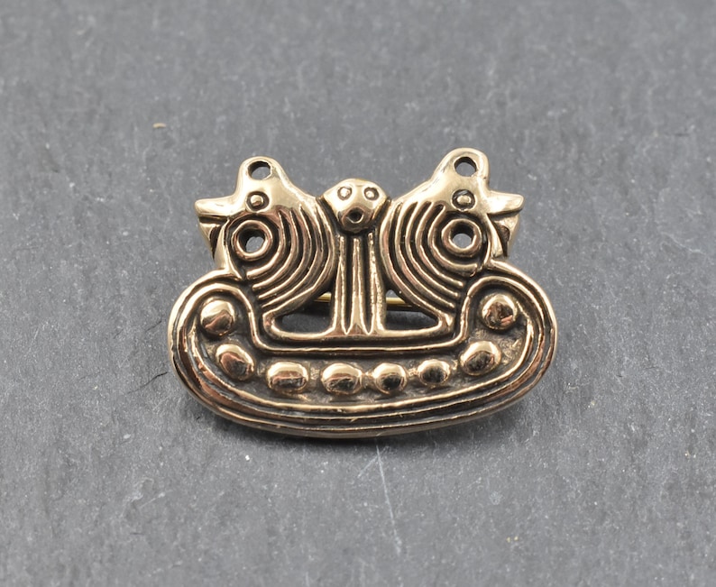 Eine goldfarbene Brosche aus Metall mit einem Design aus zwei stilisierten Vögeln, die sich gegenüberstehen. Die Vögel sitzen auf einem bootförmigen Sockel mit einer Reihe runder Akzente.