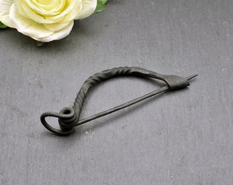 May include: A black metal brooch with a twisted wire design and a pin. The brooch is in the shape of a circle with a pointed end.