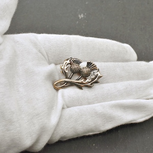Puede incluir: Un broche met&aacute;lico con un dise&ntilde;o de dos cabezas de flor de cardo con patrones texturizados en forma de rejilla en la parte superior, acompa&ntilde;adas por dos hojas estilizadas y dentadas que se curvan a lo largo de la base de los tallos.
