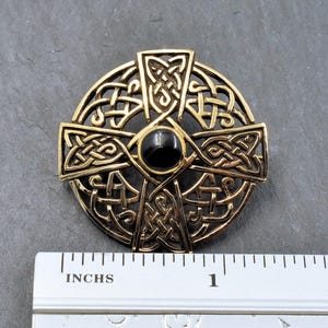 Ein goldfarbener keltischer Kreuzanhänger mit aufwendigem Knotenmuster und einem schwarzen Mittelstein. Der Anhänger hat einen Durchmesser von etwa 1,2", wie das Lineal zeigt. Das Kreuz ist kreisförmig mit einem detaillierten, durchbrochenen Muster.
