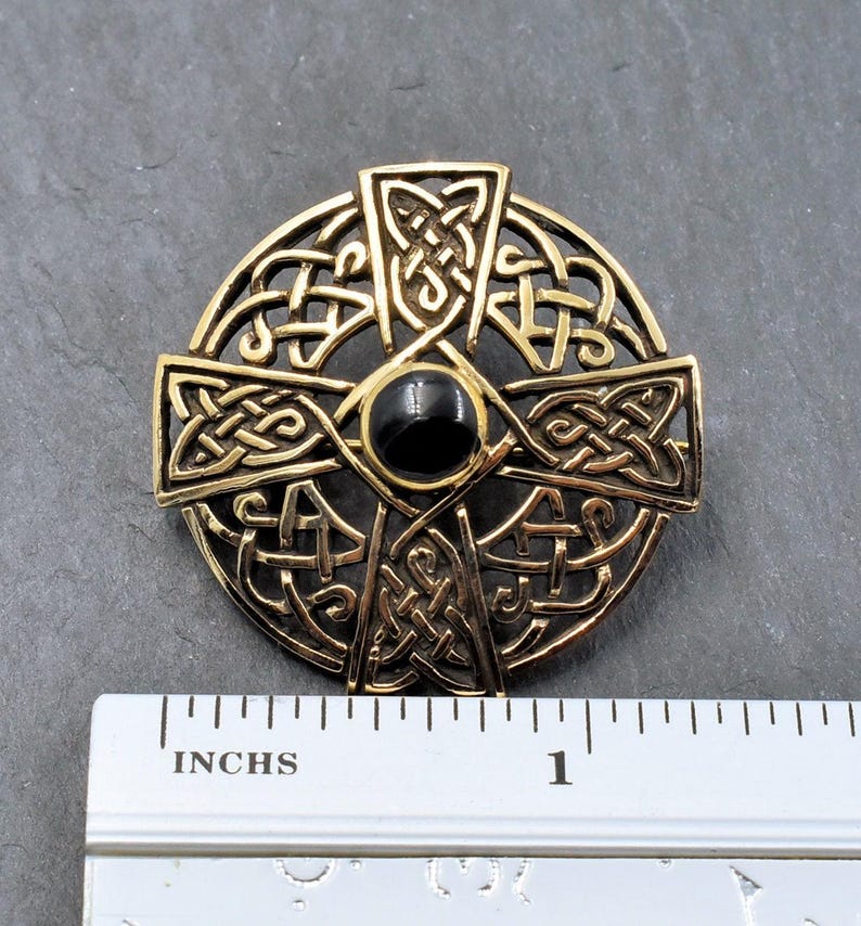 Ein goldfarbener keltischer Kreuzanhänger mit aufwendigem Knotenmuster und einem schwarzen Mittelstein. Der Anhänger hat einen Durchmesser von etwa 1,2", wie das Lineal zeigt. Das Kreuz ist kreisförmig mit einem detaillierten, durchbrochenen Muster.
