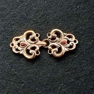 Könnte beinhalten: Ein goldfarbener Verschluss mit einem verzierten, wirbelnden Design. Der Verschluss besteht aus zwei Teilen, jeweils mit einem zentralen roten Stein und dekorativen Locken. Der Verschluss dient zum Verbinden von Stoff- oder Materialteilen.