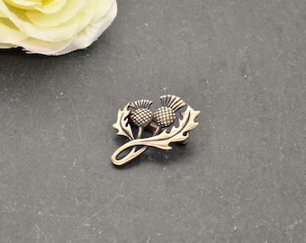 May include: A metallic brooch featuring a detailed design of two thistle flower heads with textured, rounded tops and pointed, serrated leaves extending from the base. The piece has a warm, bronze-toned finish with darker recessed areas.