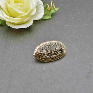 Könnte beinhalten: Eine ovale Brosche in Goldton mit einem filigranen, durchbrochenen Design. Das Design zeigt ein zentrales Kreuz mit wirbelnden Mustern und Knoten.