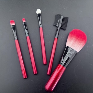 Könnte beinhalten: Satz mit fünf roten und schwarzen Make-up-Pinseln. Die Pinsel umfassen einen großen Puderpinsel, einen kleinen schrägen Pinsel, einen kleinen flachen Pinsel, einen kleinen schrägen Pinsel mit weißer Spitze und einen kleinen Kamm-Pinsel.