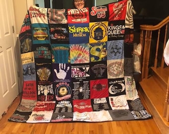 May include: A handmade quilt made from various t-shirts, featuring a patchwork design. The quilt displays a variety of colors and graphics, including text and logos from different events and organizations. The quilt is displayed indoors.