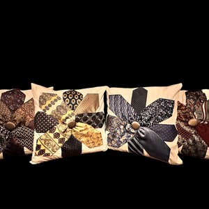 Puede incluir: Cuatro cojines decorativos con un diseño floral hecho de corbatas estampadas. Cada cojín tiene un fondo beige y un botón de madera en el centro. Las corbatas son en tonos azules, marrones y dorados.