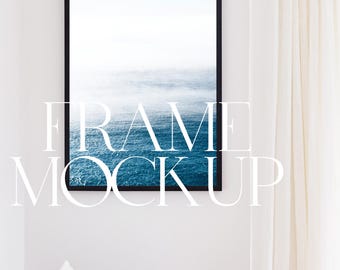 May include: A black framed picture of a blue ocean with white clouds in the sky hanging on a white wall. The wall is next to a white curtain and a wooden chair with white and beige pillows.