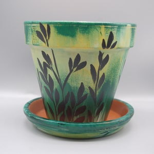 May include: A green and yellow terracotta planter with a black leafy design. The planter has a matching saucer.