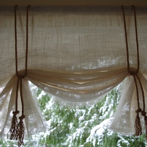 May include: White sheer curtains with a gathered, ruffled design and brown rope tiebacks with tassels. The curtains are hung on a wooden rod and are partially open, revealing a view of a snowy forest.