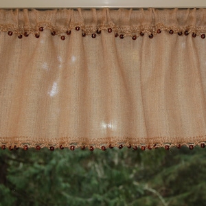 May include: A brown burlap valance with a crocheted trim and brown beads. The valance is hanging in a window with a view of green trees.