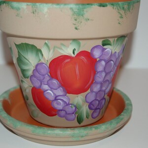 May include: A terracotta flower pot with a painted design of red apples and purple grapes. The pot has a green rim and a matching saucer.