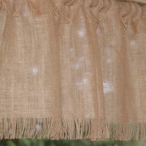 May include: A tan burlap window valance with a fringed bottom edge and a gathered top. The valance is made of a coarse, woven fabric and hangs in front of a window. The valance is a neutral color and would complement many decor styles.
