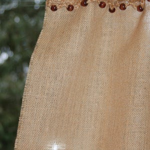 May include: A brown burlap valance with a crocheted trim and brown wooden beads. The valance is hanging on a window with a green background.