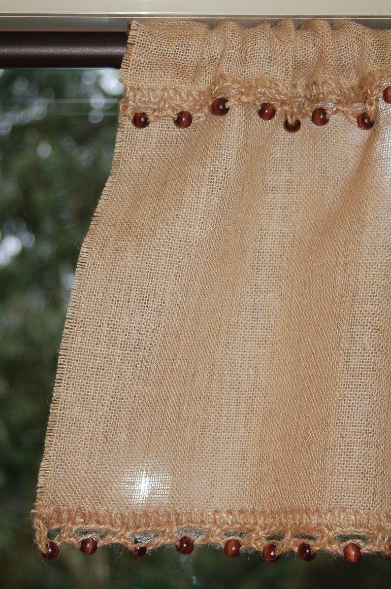 May include: A brown burlap valance with a crocheted trim and brown wooden beads. The valance is hanging on a window with a green background.