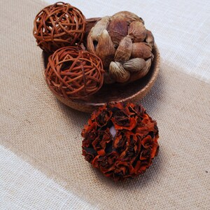K&ouml;nnte beinhalten: Eine Holzschale gef&uuml;llt mit dekorativen Naturmaterialien, darunter eine braune geflochtene Kugel, eine braune und beige geflochtene Kugel, eine braune und beige stachelige Kugel und eine braune und orangefarbene bl&uuml;tenf&ouml;rmige Kugel.