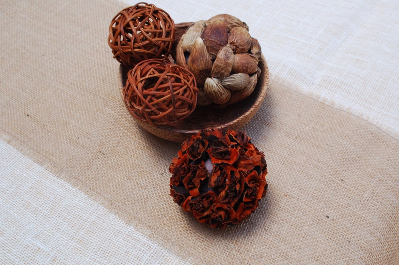 K&ouml;nnte beinhalten: Eine Holzschale gef&uuml;llt mit dekorativen Naturmaterialien, darunter eine braune geflochtene Kugel, eine braune und beige geflochtene Kugel, eine braune und beige stachelige Kugel und eine braune und orangefarbene bl&uuml;tenf&ouml;rmige Kugel.