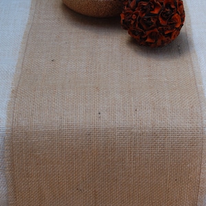 K&ouml;nnte beinhalten: Ein brauner Tischl&auml;ufer aus Jute mit einer nat&uuml;rlichen, gewebten Textur. Der L&auml;ufer liegt auf einer wei&szlig;en Tischdecke und hat eine Schale mit Trockenfr&uuml;chten und eine braune Dekorkugel darauf.