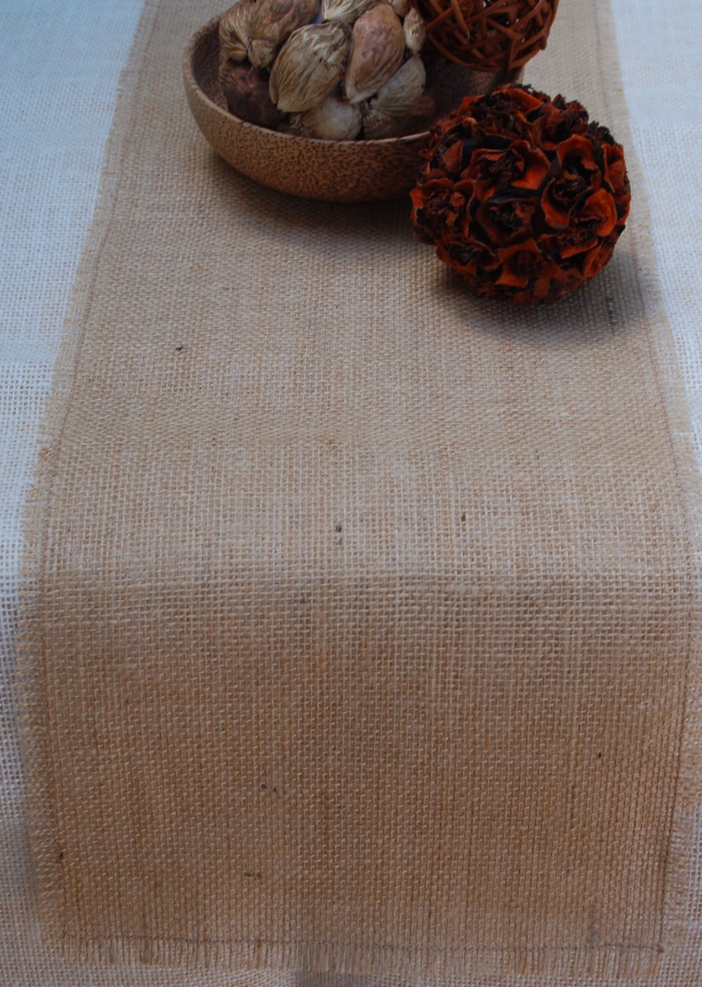 K&ouml;nnte beinhalten: Ein brauner Tischl&auml;ufer aus Jute mit einer nat&uuml;rlichen, gewebten Textur. Der L&auml;ufer liegt auf einer wei&szlig;en Tischdecke und hat eine Schale mit Trockenfr&uuml;chten und eine braune Dekorkugel darauf.