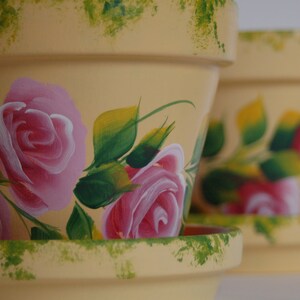 May include: Two yellow terracotta flower pots with hand-painted pink roses and green leaves. The pots have a rustic, vintage look.